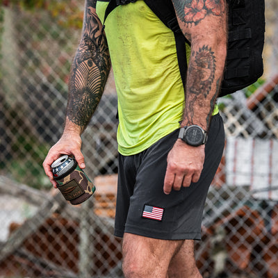 A man with tattoos holds the GORUCK Ballistic Beer Jacket, a camo can cooler ideal for keeping his cold beer chilled. He sports a yellow shirt, black shorts with a flag design, and carries a durable black backpack crafted from 1000D CORDURA.