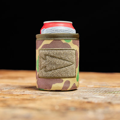 A GORUCK Ballistic Beer Jacket cradles a cold can on a wooden surface, set against a black backdrop.