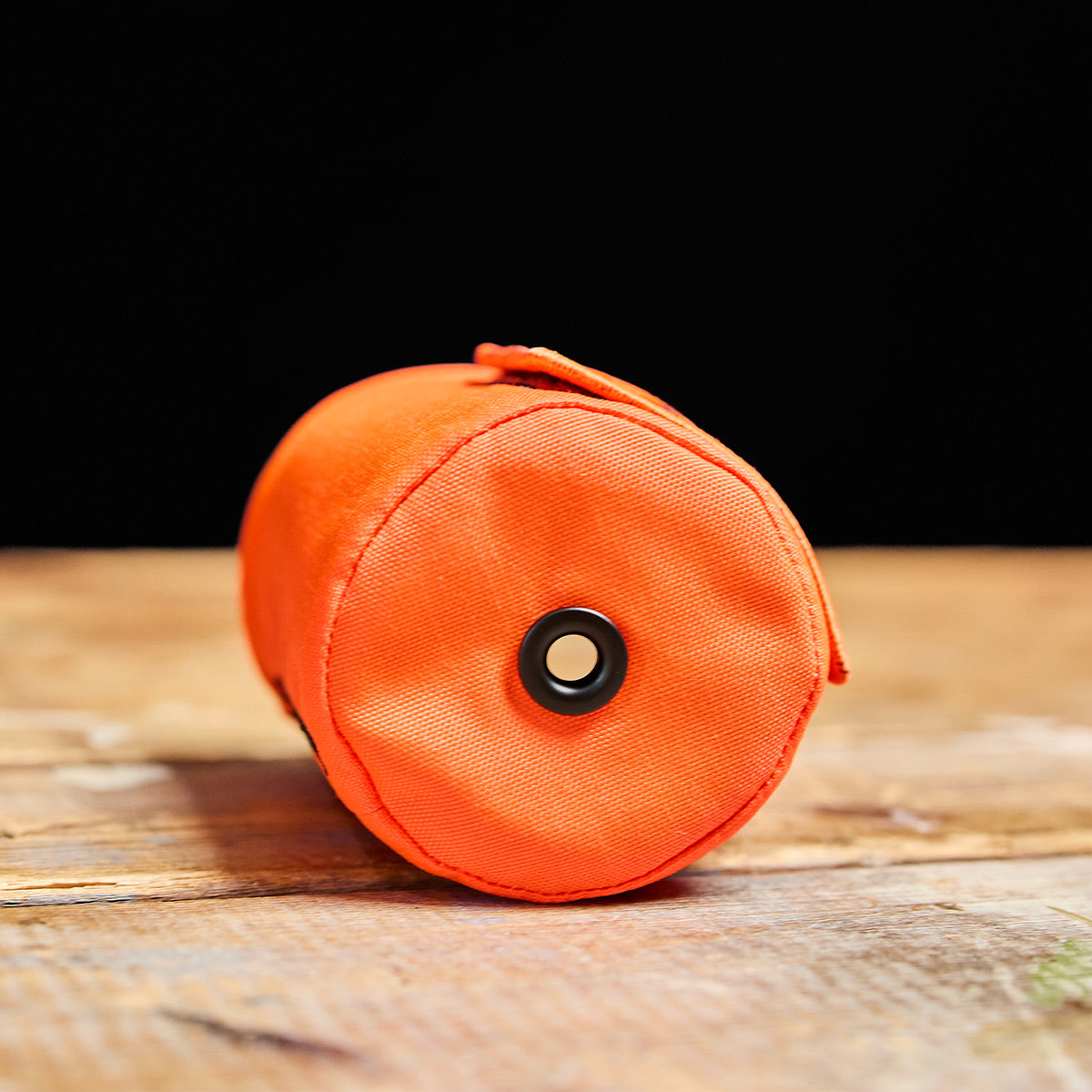 The Ballistic Beer Jacket by GORUCK, crafted from durable 1000D CORDURA in a vibrant orange hue, is displayed on a wooden surface with a black eyelet at the center—ideal for keeping your beer cold.