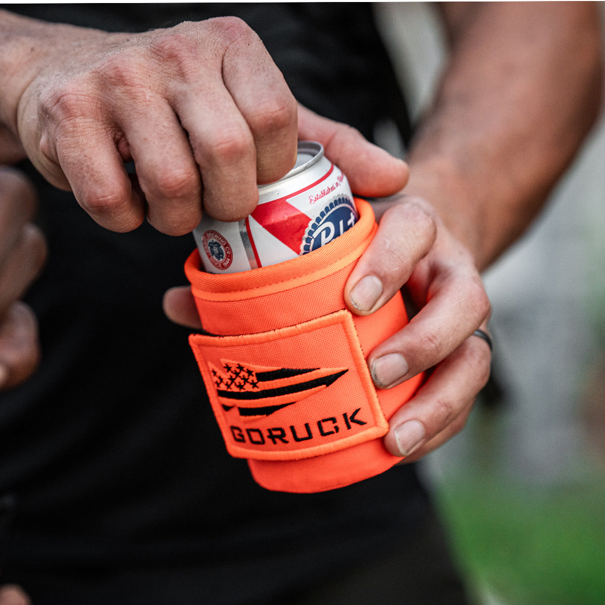 A person holds a can snugly wrapped in an orange Ballistic Beer Jacket by GORUCK, crafted from durable 1000D CORDURA.