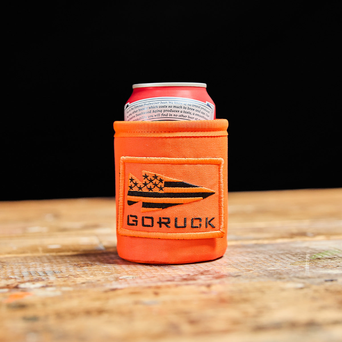 An orange Ballistic Beer Jacket by GORUCK, made from resilient 1000D CORDURA, holds a cold beer on a wooden table against a black background.