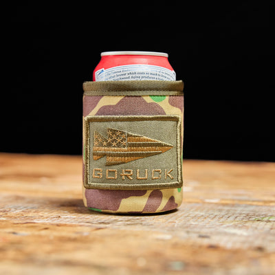 The GORUCK Ballistic Beer Jacket, crafted with durable 1000D CORDURA fabric, snugly holds a soda can on a wooden surface. Ideal for keeping your cold beer chilled, this can cooler perfectly combines ruggedness with style, ensuring that every sip you take remains refreshingly cool.