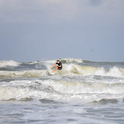 A person dressed in a black wetsuit is carving through ocean waves beneath a cloudy sky. The white, frothy swells intensify the dynamic scene as their 300 Boardshorts by GORUCK flutter in the wind.