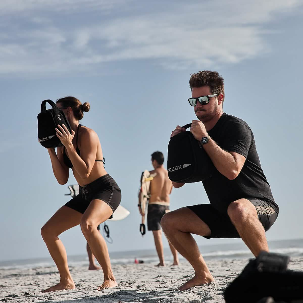 Two individuals wearing 300 Boardshorts by GORUCK perform squats on a sandy beach, holding weighted bags labeled RUCK. A man in the background seems to be resting with his surfboard, while the ocean and a cloudy sky stretch out in the distance.