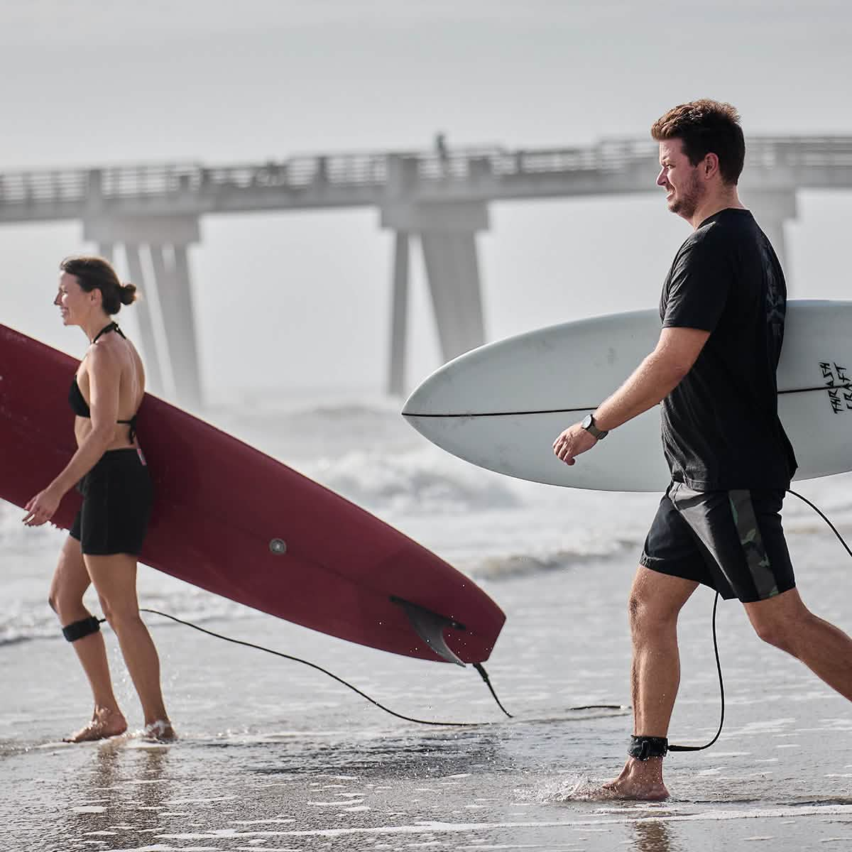 Two people stroll along a beach with GORUCK surfboards. The woman on the left carries a red board, and the man on the right holds a white one. A pier stretches into the ocean in the background. Proudly made in USA, both wear wetsuits and ankle surf leashes as they head for adventure.