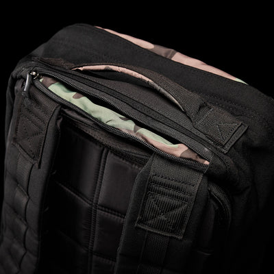 A close-up of the GR1 Birdwell backpack by GORUCK features padded straps and grid stitching, highlighting its SurfNyl™️ fabric. A partially open zipper reveals a camouflage pattern inside. This backpack, representing American craftsmanship, is set against a dark background.