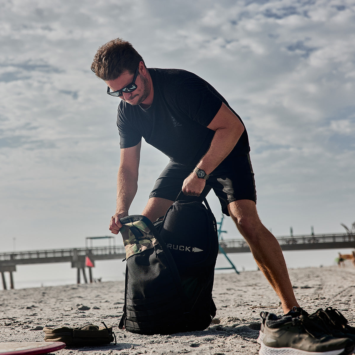 A man in sunglasses and a black outfit stands on a sandy beach, expertly packing the GR1 Birdwell by GORUCK into a black bag crafted with American craftsmanship. A pier extends over the ocean in the background under a partly cloudy sky.