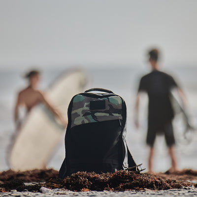 A GR1 Birdwell backpack from GORUCK, featuring a camouflage design, rests on the beach as two surfers with blurred surfboards prepare to enter the ocean. This scene, showcasing American craftsmanship, embodies an adventurous outdoor spirit.