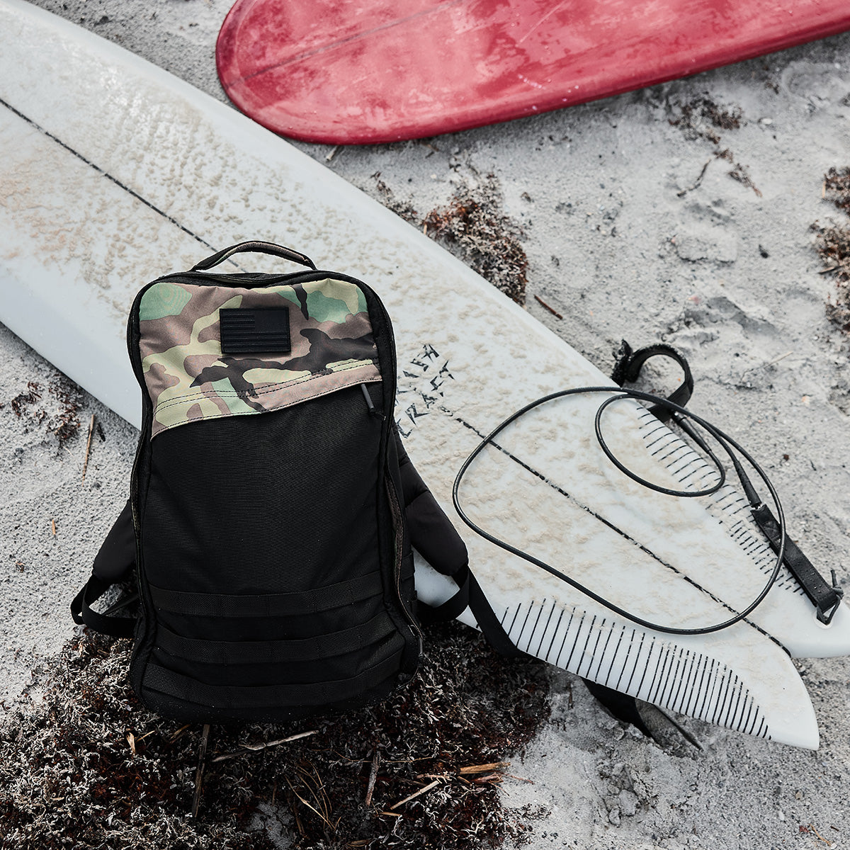 A camouflage backpack rests on the sandy beach next to a GR1 Birdwell surfboard by GORUCK, with a red surfboard partially visible in the background. A SurfNyl™️ leash is coiled beside the backpack as seaweed and sand are scattered around, highlighting American craftsmanship amidst nature's beauty.