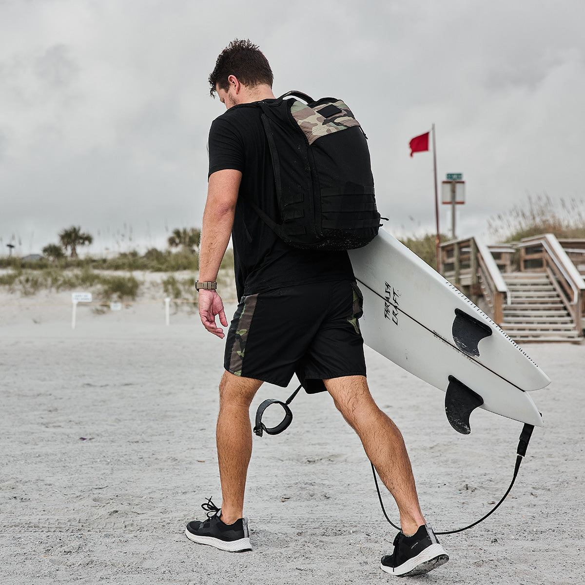 A person dressed in black athletic attire and carrying a camouflage GORUCK GR1 Birdwell backpack walks along the sandy beach. They hold a SurfNyl™️ surfboard leash, moving toward the ocean under a cloudy sky. In the background, a red flag and wooden steps are visible.