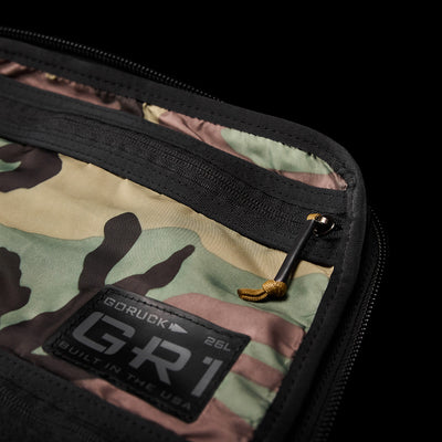 Close-up of a camo-patterned GR1 Birdwell backpack pocket with a black zipper partly open. A small black tag reads GORUCK, highlighting American craftsmanship. A yellow zipper pull complements the design, crafted with durable SurfNyl™ fabric for lasting wear.