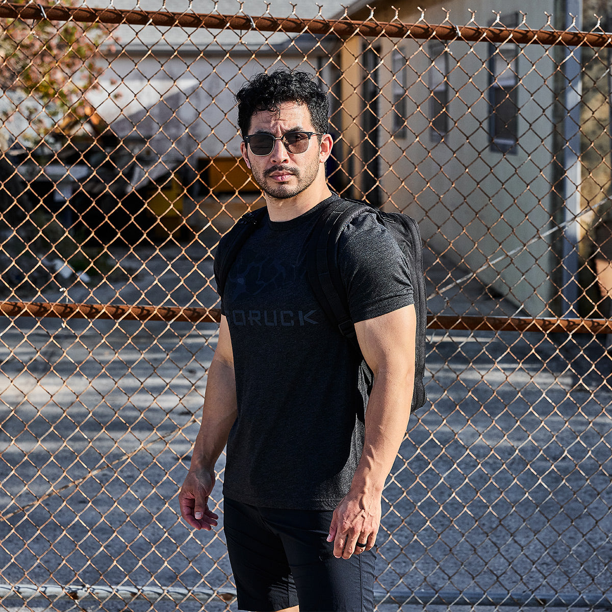 A man with sunglasses stands confidently in front of a chain-link fence, wearing a GORUCK Spearhead Tee - Tri-Blend by vendor-unknown and carrying a backpack. The background features a blurred residential building and a sunny sky.