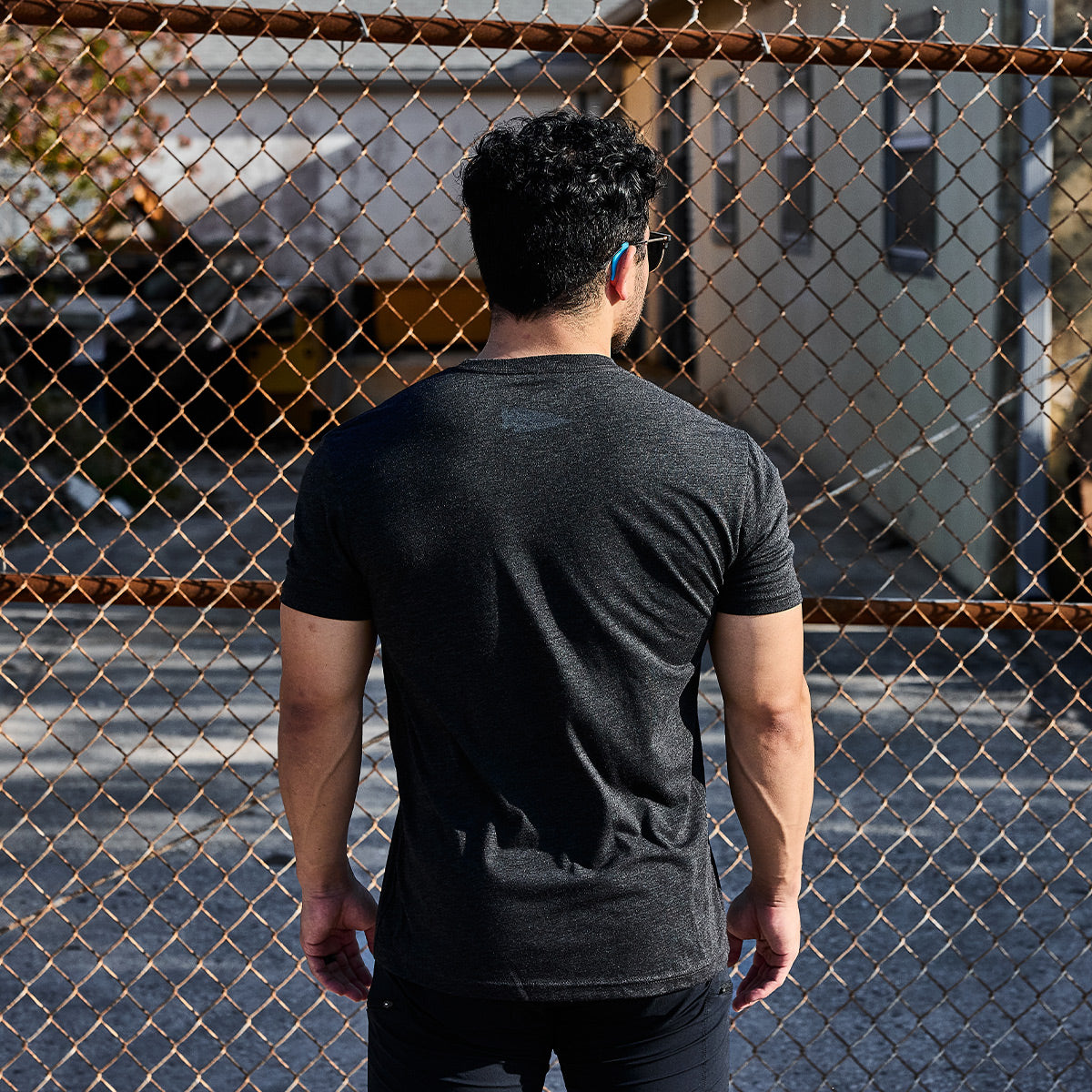 Wearing the GORUCK Spearhead Tee - Tri-Blend, a person with short hair stands facing a chain-link fence outdoors. The background reveals a building framed by tree branches, evoking the resilience and readiness inspired by Special Forces roots.