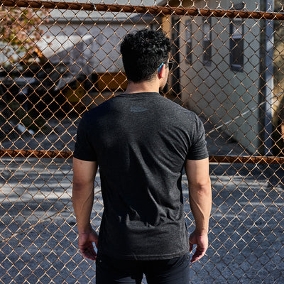 Wearing the GORUCK Spearhead Tee - Tri-Blend, a person with short hair stands facing a chain-link fence outdoors. The background reveals a building framed by tree branches, evoking the resilience and readiness inspired by Special Forces roots.