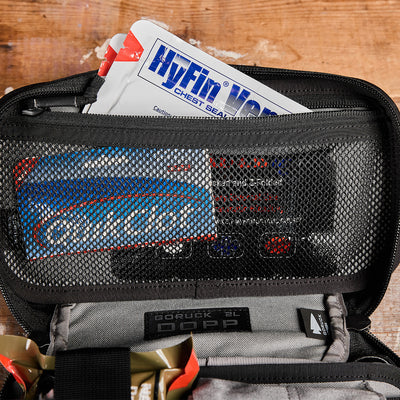 A GORUCK Dopp Kit featuring a HyFin vent chest seal, bandages, and medical tape visible through a mesh pocket is seen labeled as prepared for emergencies, resting on a wooden surface.