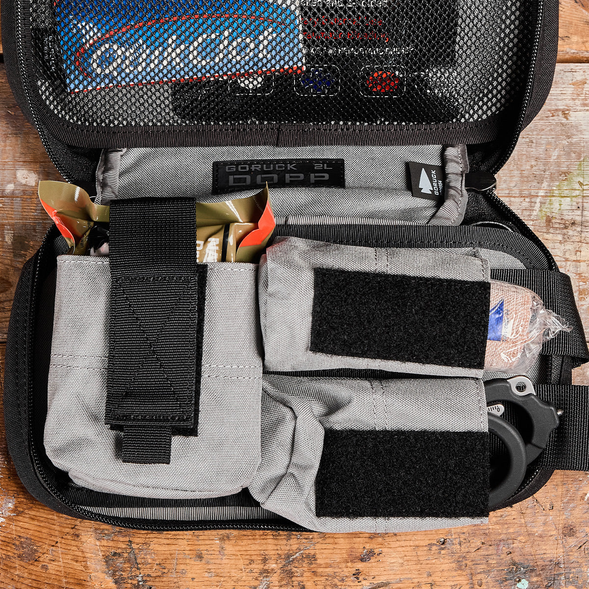 A GORUCK Dopp Kit rests on a wooden surface, showcasing MOLLE webbing and organized compartments filled with items such as tool pouches and Velcro straps. Inside, a visible pack of O'Keeffe's skin care product is neatly tucked away. The mesh cover is partially open, revealing the carefully arranged contents.