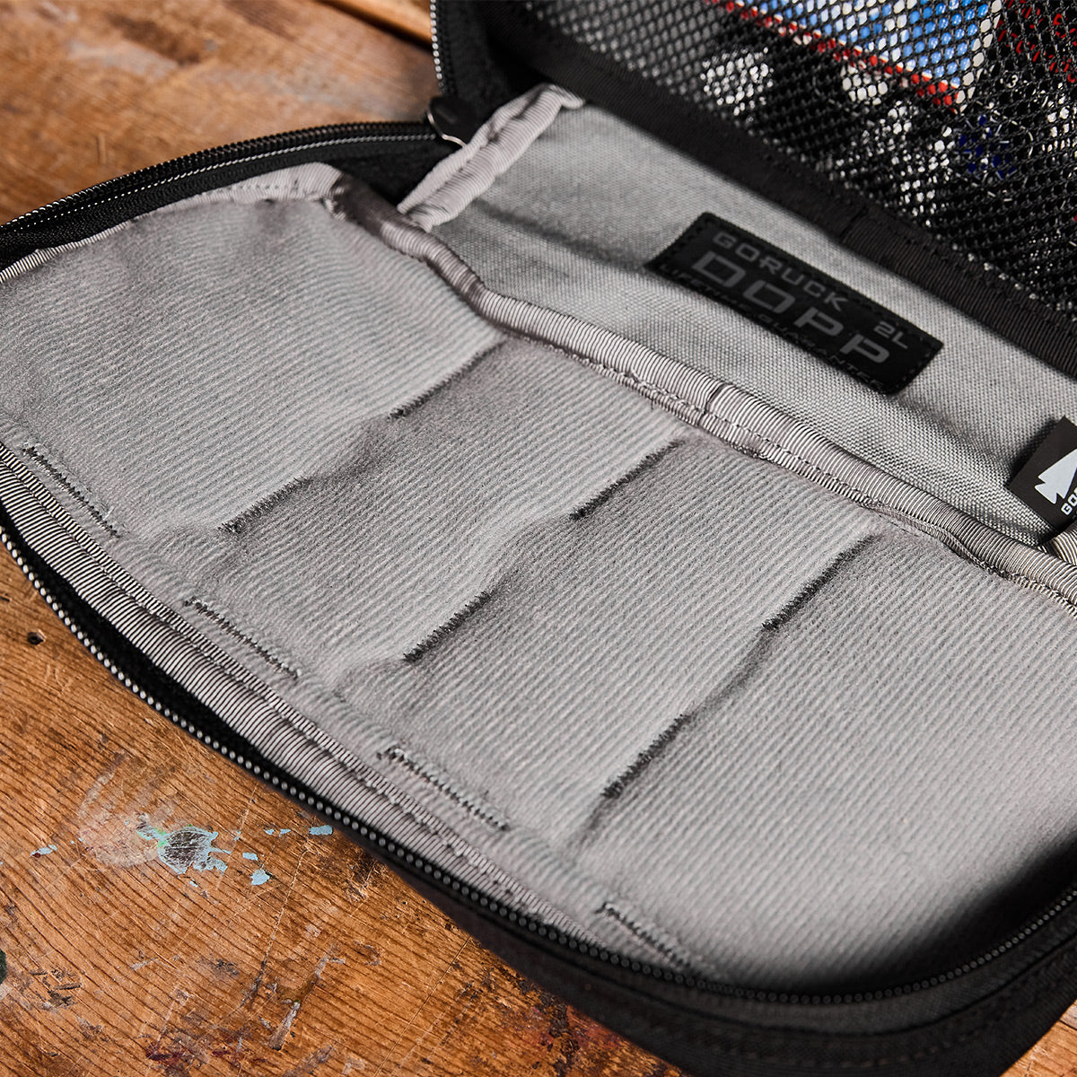 A close-up of the GORUCK Dopp Kit shows its gray fabric interior, featuring multiple compartments and a zippered mesh pocket. Designed with MOLLE webbing for added versatility, this 21L pouch is placed on a paint-splattered wooden surface.