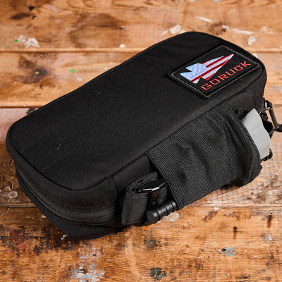 A black Dopp Kit by GORUCK, with a rectangular shape and MOLLE webbing, features an American flag patch and the GORUCK text. It rests on a rustic wooden surface, boasting several compartments and straps for securing contents.