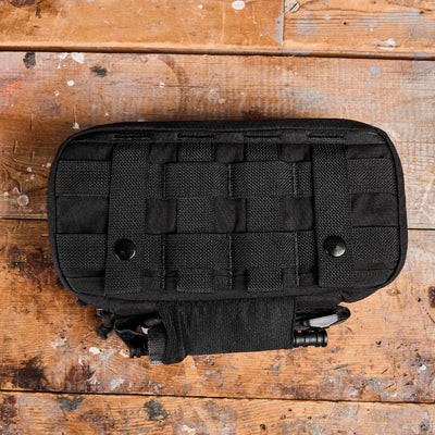 A black GORUCK Dopp Kit featuring MOLLE webbing and IFAK capabilities rests on a worn, paint-splattered wooden surface. This compact, rugged accessory is ideal for outdoor or tactical use.