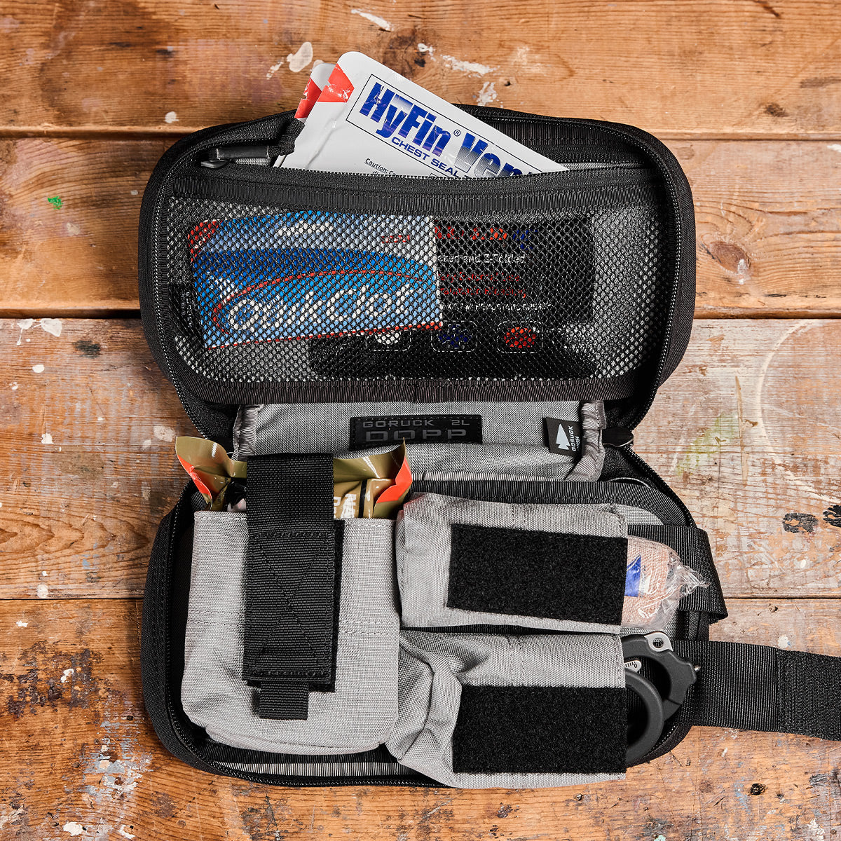 A GORUCK Dopp Kit, open on a wooden surface, reveals its contents similarly to an IFAK, with bandages, medical shears, and other emergency supplies neatly organized in compartments. A HyFin Vent package is visible at the top.