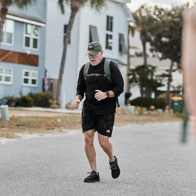 A man, donning a green cap with a black shirt and shorts, is sprinting along a paved street carrying a backpack made of CORDURA Ballistic Nylon. In this residential neighborhood adorned with houses and palm trees, his GORUCK Men's Ballistic Trainers in Black + Charcoal provide 3X stability as he navigates the area.