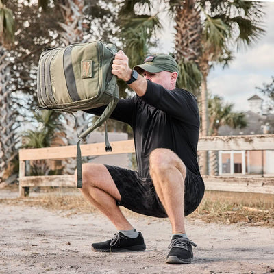 A person wearing a green cap and black athletic outfit is performing a squat with a green backpack made of durable CORDURA Ballistic Nylon. They're outdoors, with palm trees and a wooden fence in the background, demonstrating their strength in GORUCK's Men's Ballistic Trainers - Black + Black + Charcoal W/ Black Reflective Spearhead for maximum 3X Stability.