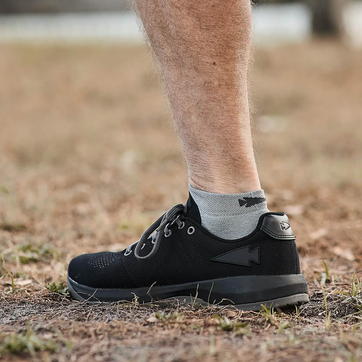 A side view captures a person's leg wearing GORUCK's Men's Ballistic Trainers - Black + Black + Charcoal with Black Reflective Spearhead, made from CORDURA Ballistic Nylon and paired with gray socks, standing on a dry, grassy surface. Designed for functional fitness with an emphasis on 3X Stability, the footwear stands out against the blurred background.