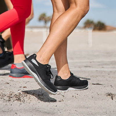 A group of people jogs along a sandy beach, concentrating on their legs and footwear. The leader is wearing GORUCK Women's Ballistic Trainers in Black and Glacier Grey, featuring Silver Reflective Spearhead accents crafted from durable CORDURA Ballistic Nylon. The person behind dons coral leggings paired with functional fitness shoes offering 3X Support. Palm trees sway gently in the background.