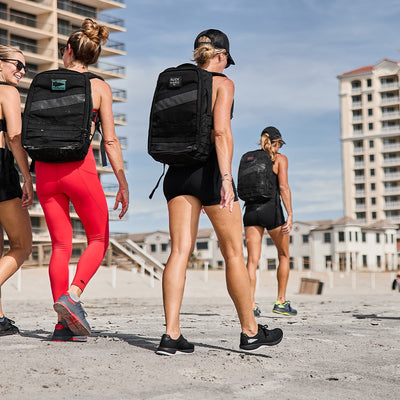 Four women wearing athletic clothing and backpacks walk along a sandy beach, highlighting their Women's Ballistic Trainers in Black and Glacier Grey with Silver Reflective Spearhead from GORUCK. They stroll near high-rise buildings under a blue sky, embodying the essence of 3X Support for an active lifestyle.