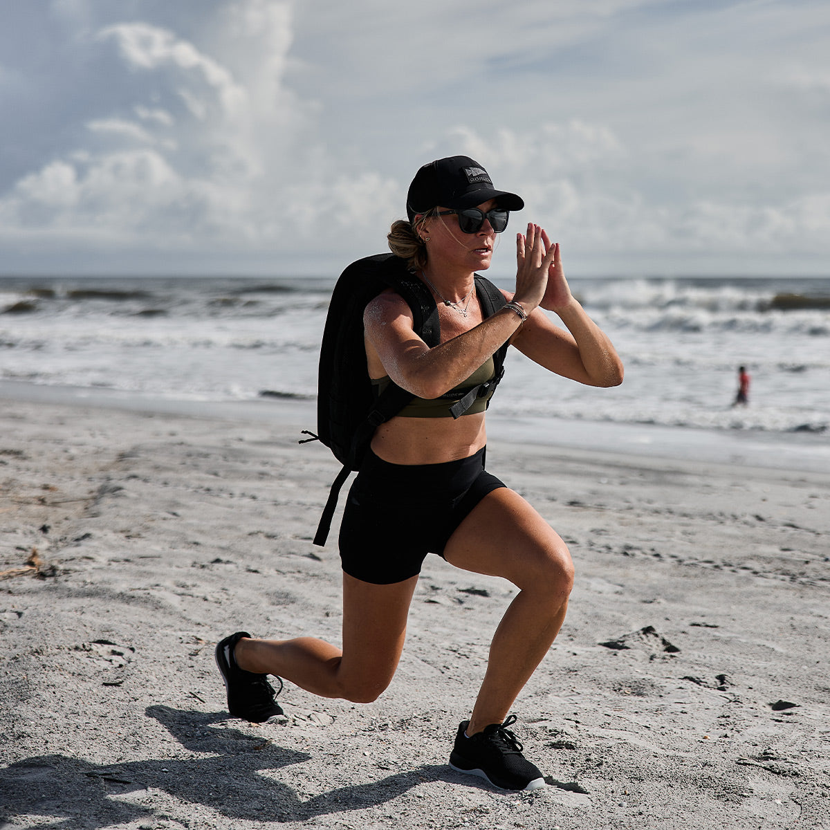 A person exercises on a sandy beach, wearing a backpack, cap, sports bra, and shorts crafted from CORDURA Ballistic Nylon. They showcase GORUCK's Women's Ballistic Trainers in Black + Glacier Grey with Silver Reflective Spearhead as they lunge with hands together, ocean waves rolling in the background under a cloudy sky.