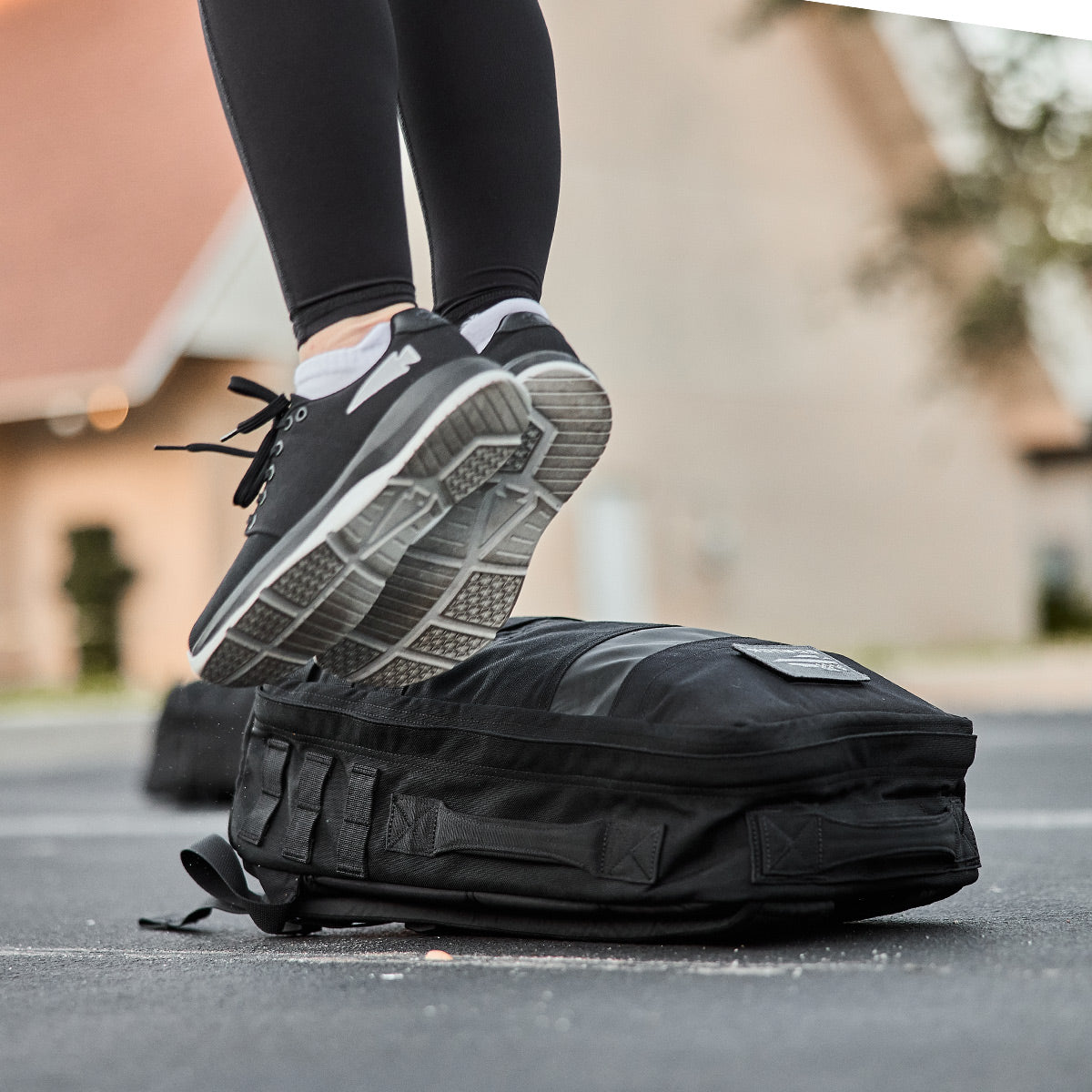 A person steps on a black backpack outdoors, dressed in black leggings and wearing GORUCK Women's Ballistic Trainers in Black and Glacier Grey with Silver Reflective Spearhead accents. The shoes are designed with CORDURA Ballistic Nylon for enhanced durability, while the blurred buildings and trees in the background suggest a suburban or campus environment.