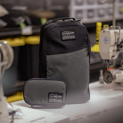 On a workbench with sewing machines in the background, a GORUCK Bullet Heritage - Built by SCARS backpack, along with a matching pouch in black and gray, is displayed. The backpack prominently features its Built by SCARS patch.