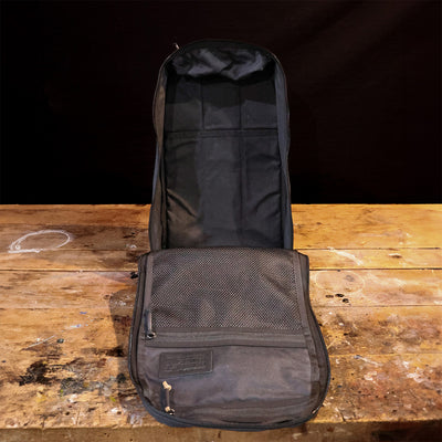 A black open case sits on a worn wooden table, with a mesh pocket on its lid. The case appears empty, evoking the nostalgic charm of a GORUCK Bullet Heritage - Built by SCARS piece, set against a dark background.