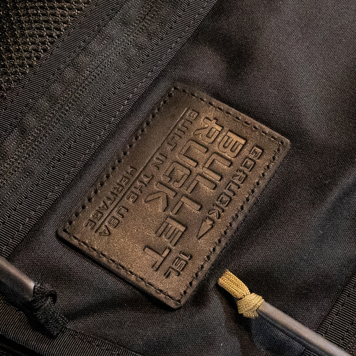 Close-up of a black fabric bag featuring a brown leather patch embossed with the text: Bullet Heritage - Built by SCARS. This retro canvas rucksack from GORUCK includes a woven mesh section and a cord with tan and black detail, exuding a classic yet functional vibe.