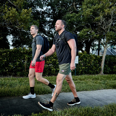 Two men walk on a sidewalk surrounded by grass and trees, wearing casual workout attire crafted from quick-drying performance fabric. Backpacks rest on their shoulders as one sports a gray GORUCK Men’s Performance Tee - ToughMesh with red shorts, and the other wears a black featherweight training shirt paired with green shorts.