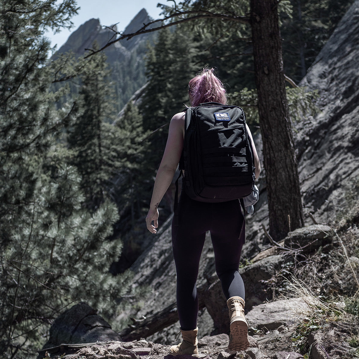 A person with pink hair, dressed in black hiking gear and sturdy boots, navigates a rocky trail through a forested mountain region. With tall trees and rugged peaks providing a stunning backdrop, they stay comfortable on their journey thanks to GORUCK's Merino Challenge Socks - Crew, renowned for comfort and durability.