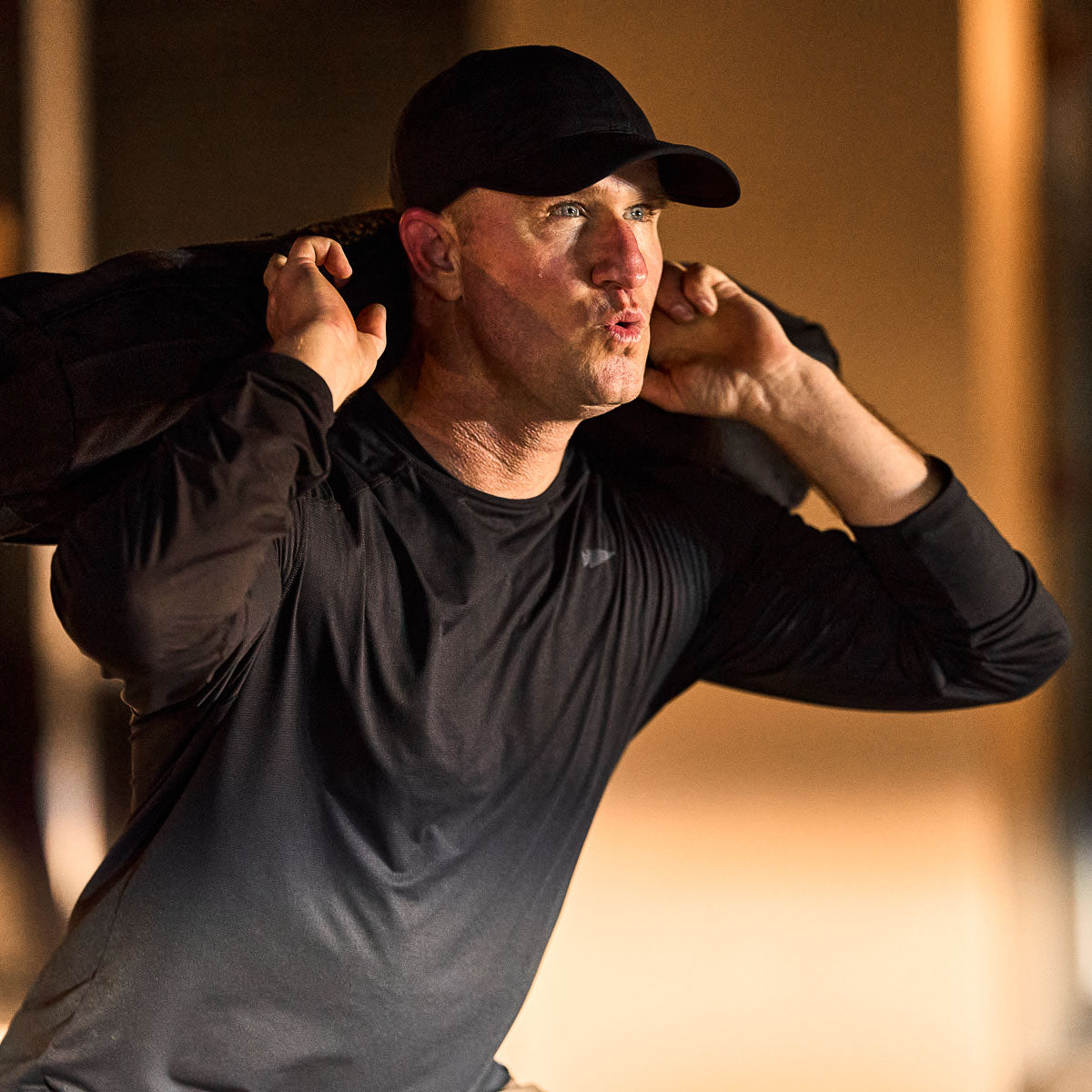 A man in a black Performance Running Hat - ToughMesh, made from Italian fabric, lifts weights with a focused expression under the soft glow of warm light.