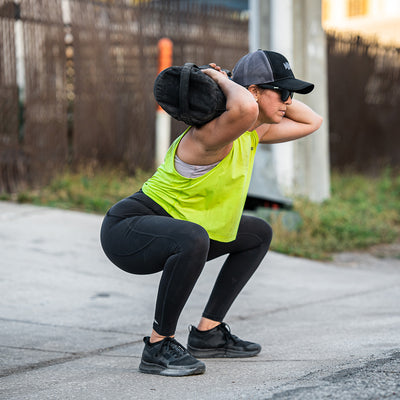 Wearing the Women's Rough Runner - Blackout by GORUCK, along with black leggings and a cap shading their eyes, a fitness enthusiast tackles a squat using a weighted sandbag on their shoulders. The outdoor setting evokes memories of Rough Runner events, set on a paved area with a fence distantly standing guard.