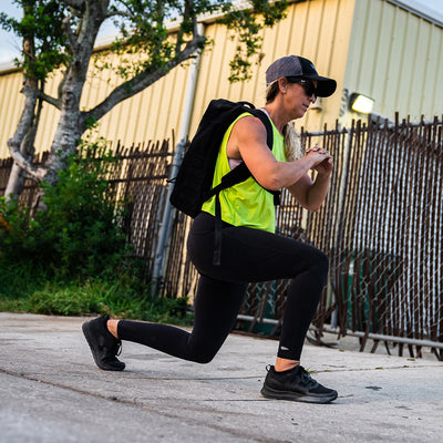 Wearing a neon green tank top, black leggings, and a cap, a person demonstrates a lunge while carrying the Women's Rough Runner - Blackout backpack by GORUCK. They are exercising outdoors on a paved surface with a chain-link fence and yellow building in the background.