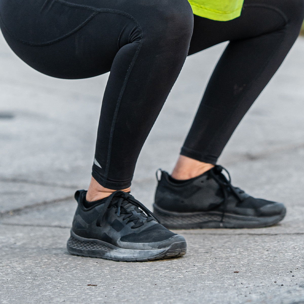 A person squatting outdoors wearing the GORUCK Women's Rough Runner - Blackout, which includes black leggings and black sneakers featuring an EVA midsole, stands on the concrete ground. Only the lower half of the body is visible, embodying the spirit of a rough runner ready for action.