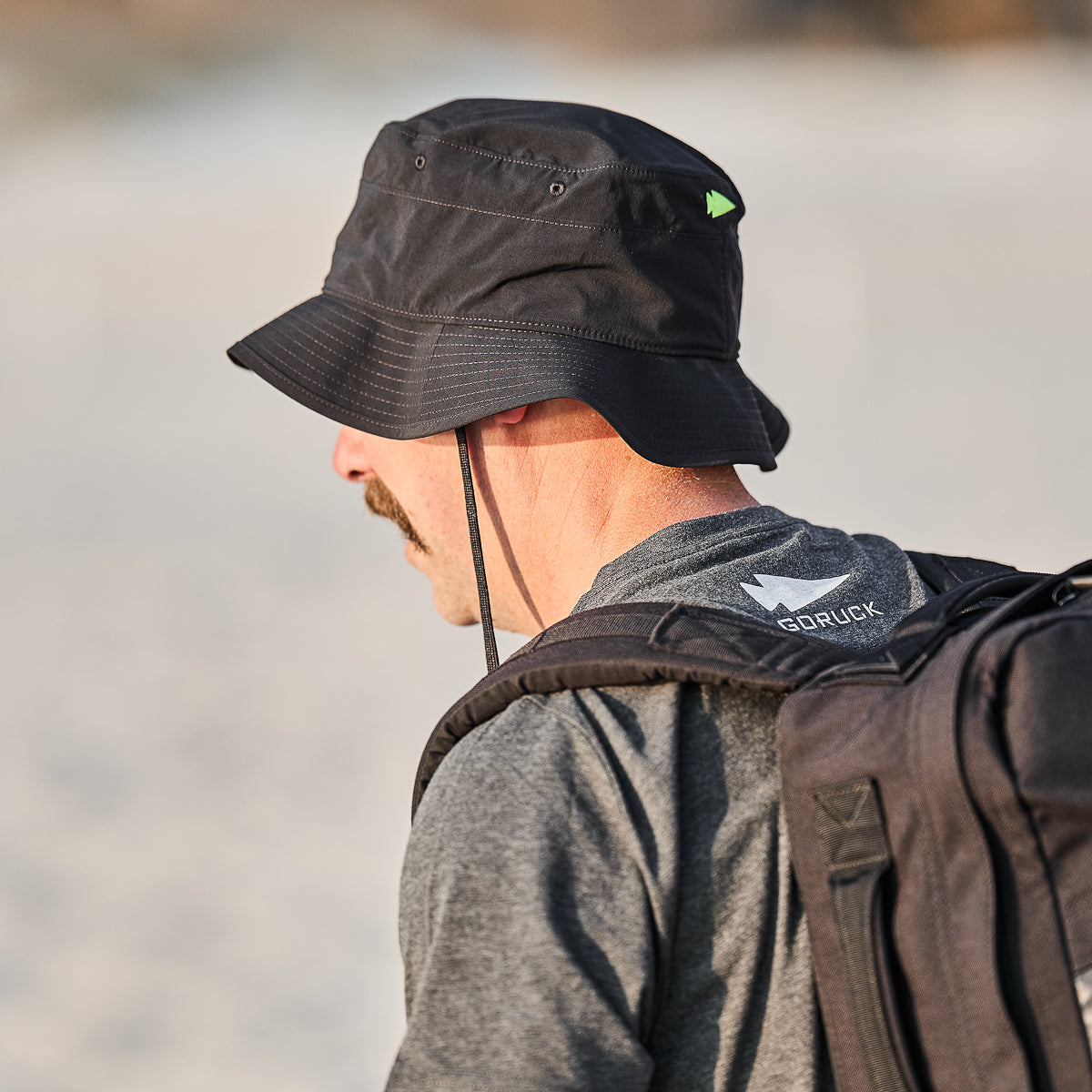 A person wearing a black Boonie Hat - Slick - ToughDry and a backpack, with "GORUCK" on their shirt, strolls along a sandy beach, enjoying protection from the elements.