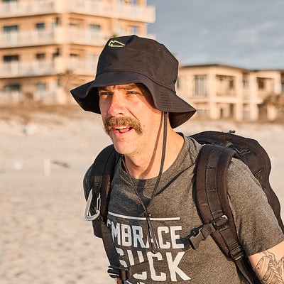 A man wearing a Boonie Hat - Slick - ToughDry and a gray shirt smiles on the beach with a backpack and mustache, enjoying protection from the elements.