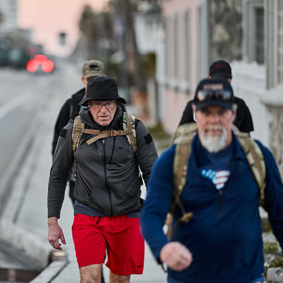 People wearing outdoor gear and backpacks, including reliable Boonie Hat - Slick - ToughDry, stroll on a sidewalk in an urban setting.