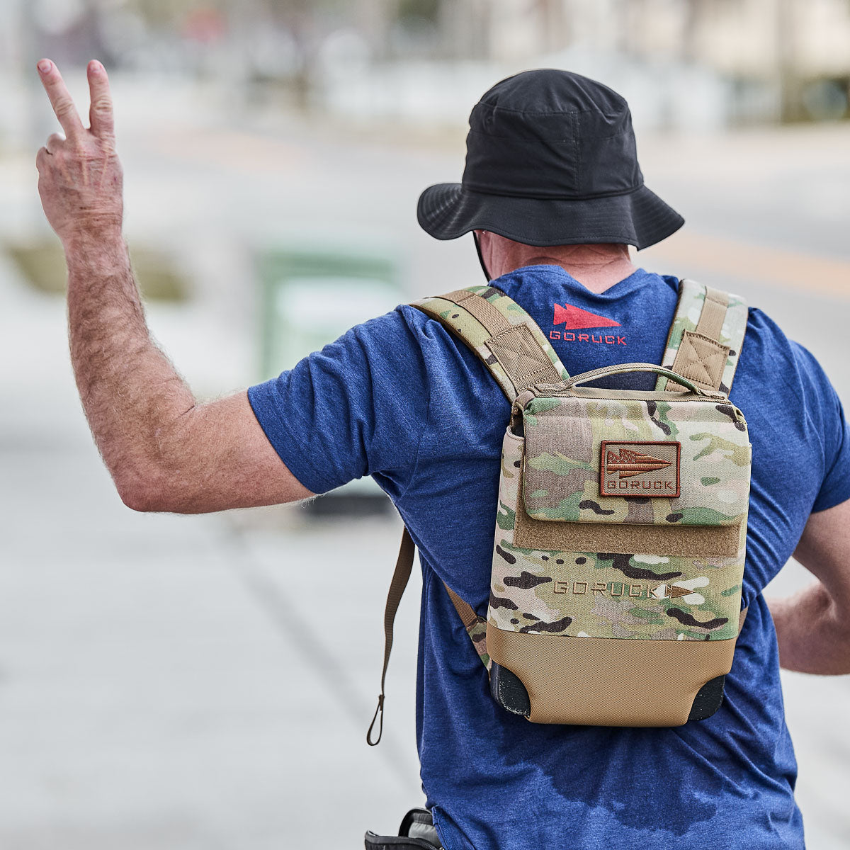 A man in a blue shirt and camo backpack strolls down a city street, flashing a peace sign. He wears a Boonie Hat - Slick - ToughDry, blending style and protection as he embraces his urban adventure.