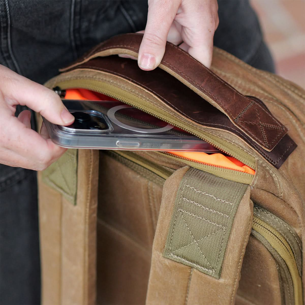 A person places a smartphone into the front pocket of the GR1 USA - Heritage rucksack by GORUCK, crafted with waxed duck canvas and adorned with green accents, effortlessly pairing style and practicality.