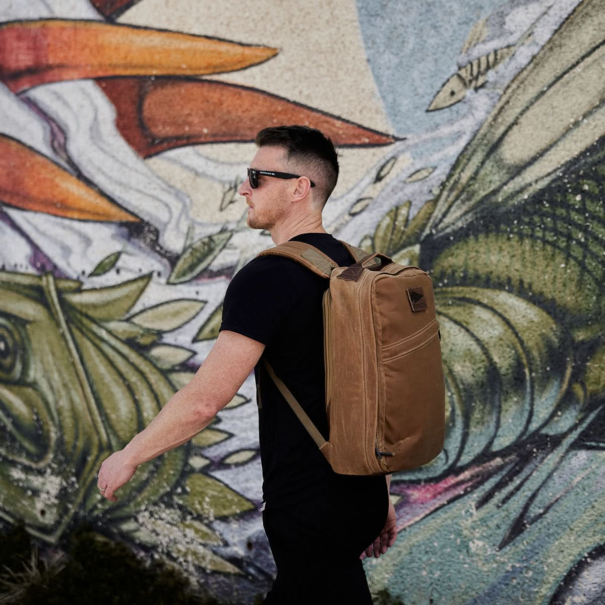 A person dressed in black carrying the GR1 USA - Heritage rucksack by GORUCK made from waxed duck canvas walks by a colorful mural.