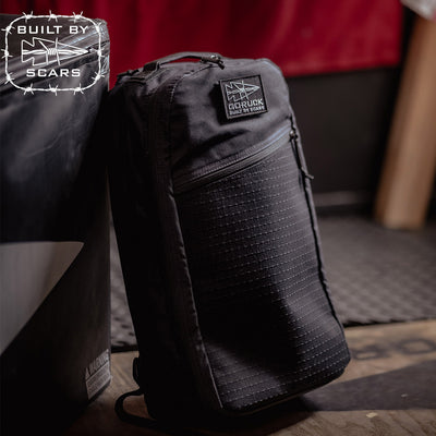 A black Bullet Heritage backpack, crafted by GORUCK's SCARS, rests against a surface. It displays a small rectangular patch that reads "Built in the USA," enhancing its rugged charm. The dimly lit background with industrial elements complements its aesthetic.