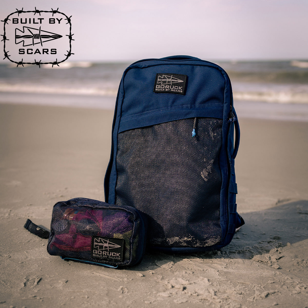 A Rucker 4.0 20L backpack in Navy with a mesh front slant and a small pouch rest on the sandy beach, framed by the ocean in the background, ideal for rucksack training. Both items prominently display the GORUCK logo, with "Built by Scars" text in the upper left corner.