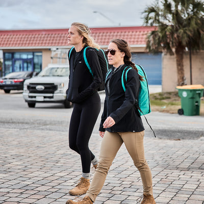 Two people stroll down a street wearing casual outdoor attire, each carrying the Bullet Ruck Classic - Cordura daypack featuring extra padded straps for optimal comfort. In the background, a truck idles beside a swaying palm tree.