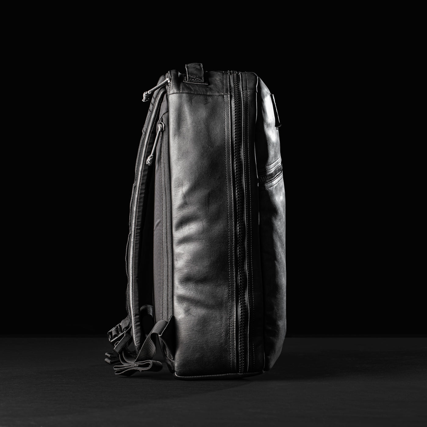 Side view of a sleek black leather Bullet Ruck x Carryology - Berserker Viking by GORUCK against a dark background. The backpack features visible zippers and adjustable straps, highlighting its modern and minimalist design.